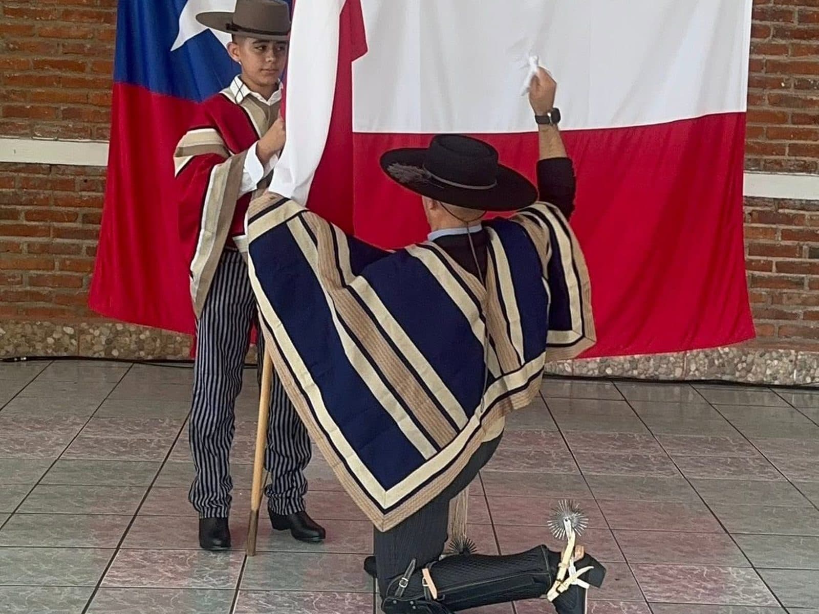 Dos bailarines de traje típico junto a la bandera: uno sostiene el asta y el otro, de manta rayada y sombrero, se arrodilla con el pañuelo en alto.
