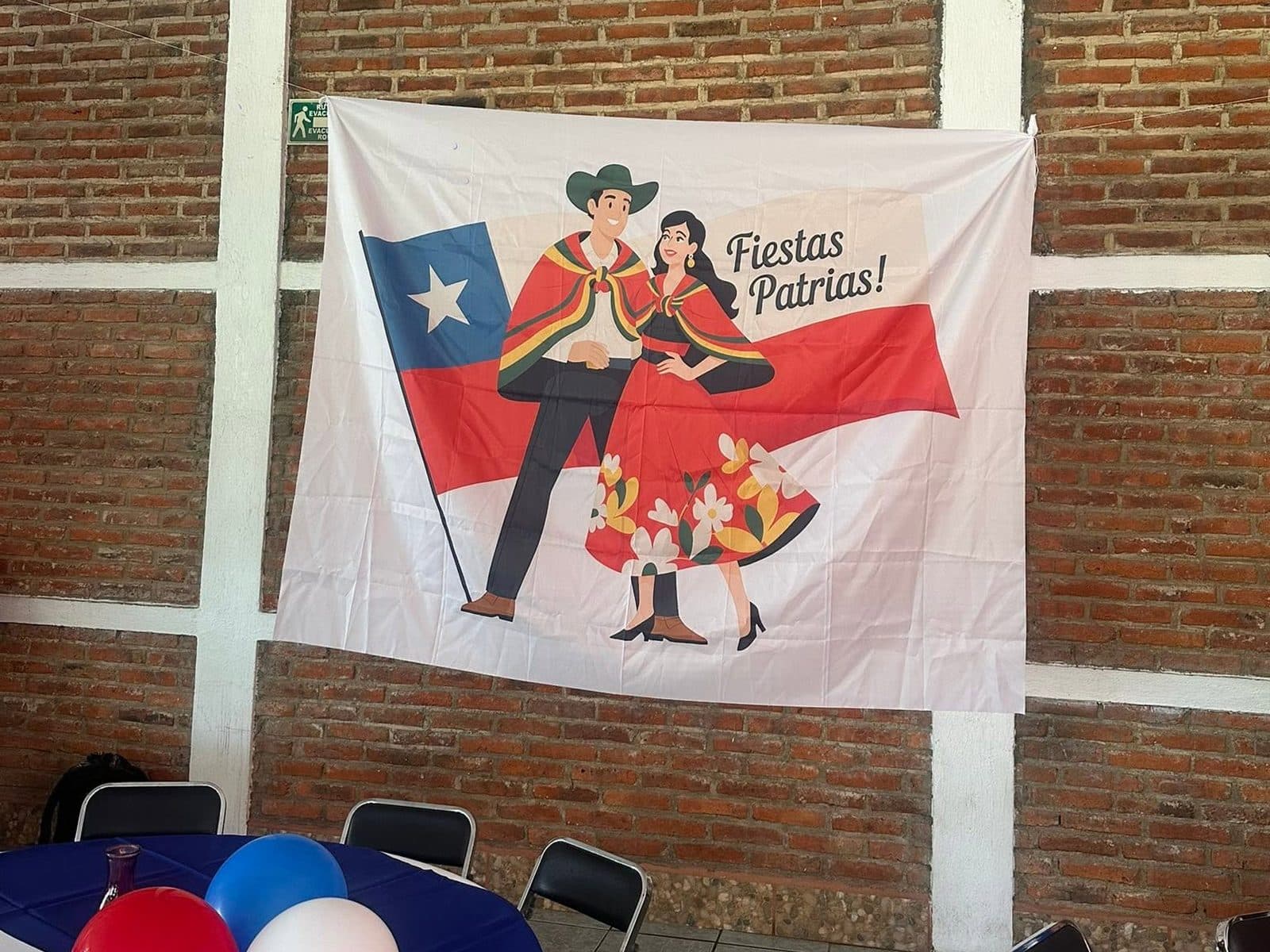 El lienzo de Fiestas Patrias colgado en la pared de ladrillo, con las mesas puestas y globos rojos, blancos y azules.