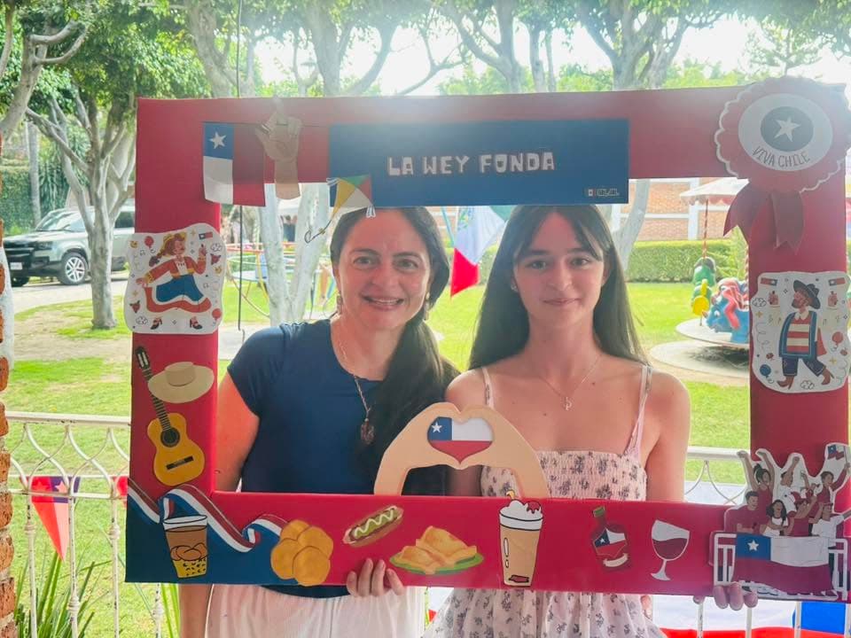 Una mamá y su hija sostienen el marco de La Wey Fonda en el jardín, decorado con banderas, una guitarra, una chupalla y dibujos de sopaipillas, completos y terremotos.