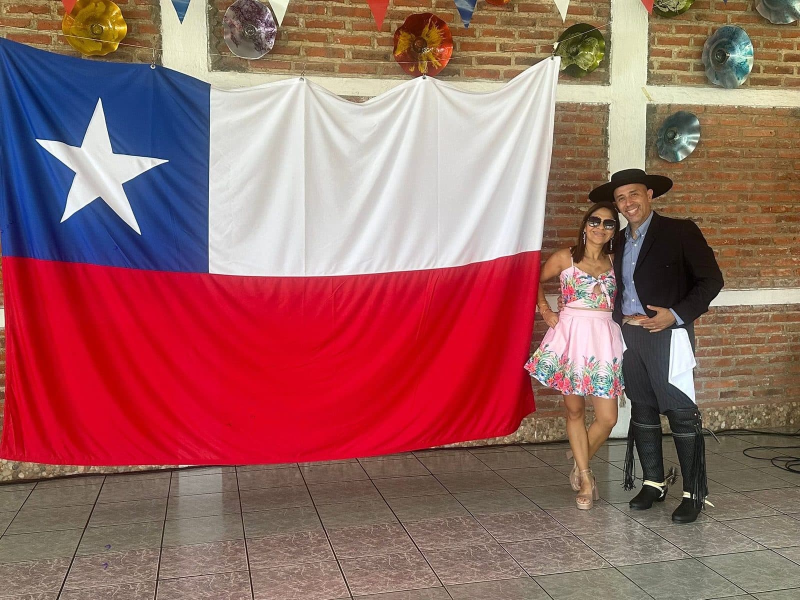 Una pareja posa frente a una bandera chilena enorme colgada en el salón: él de huaso, con sombrero de ala plana y botas con espuelas.