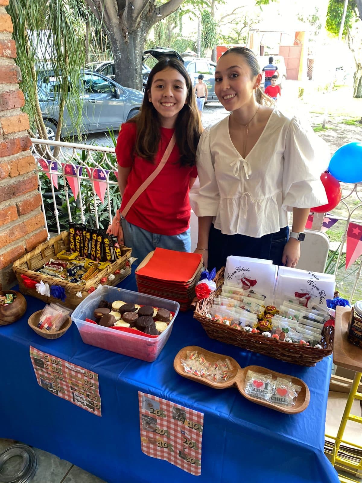 Dos jóvenes atienden el puesto de dulces chilenos: canastos con Super 8 y chocolates, alfajores, y bolsitas de calugas sobre un mantel azul con banderines.